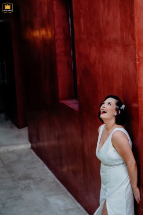 Merida, Mexico, provides a vibrant setting where a beaming bride, dressed in white, throws her head back in a moment of natural laughter while posing against a textured red wall.